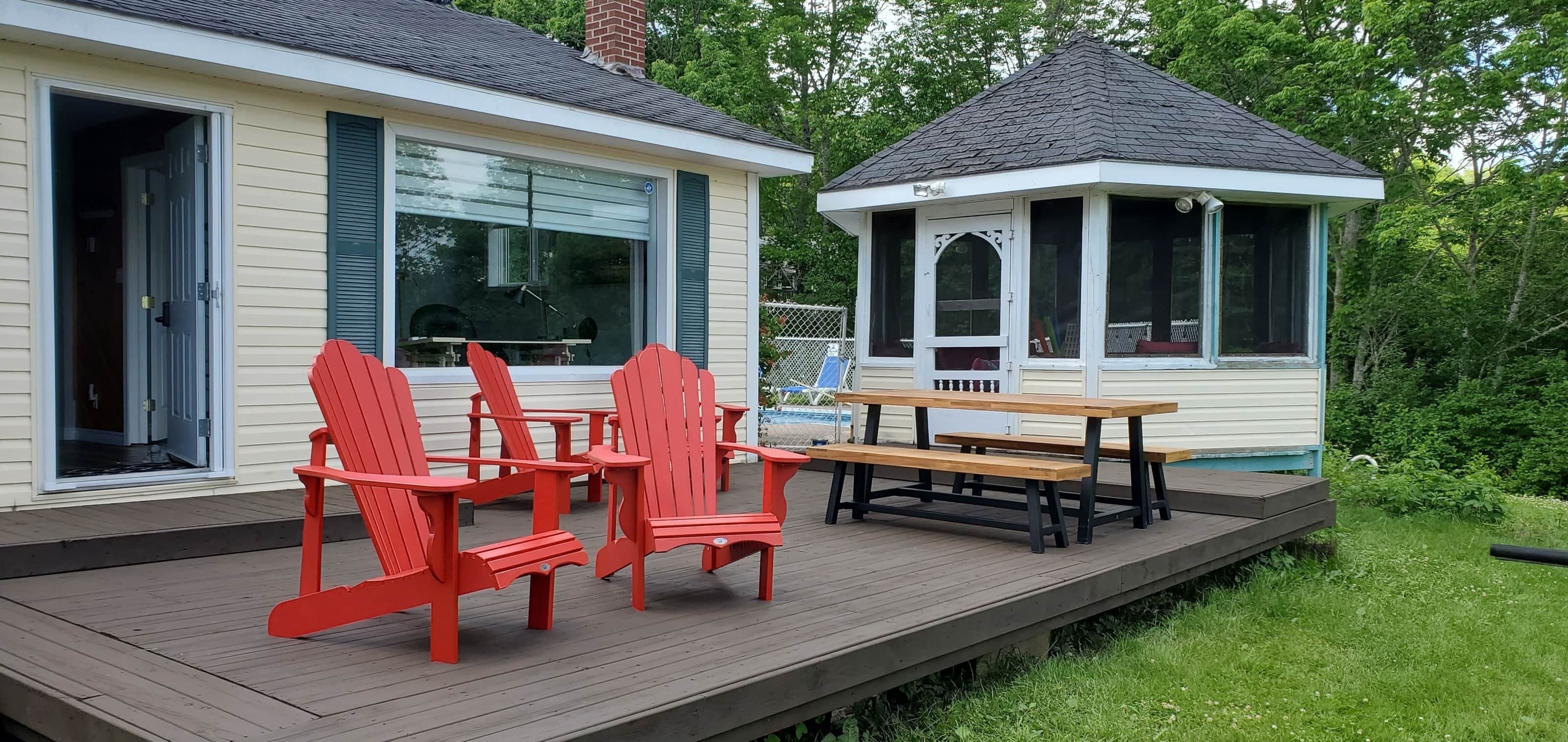 A deck with red Adirondack chairs and a picnic table, a covered gazebo, and a house with a large window and green shutters.