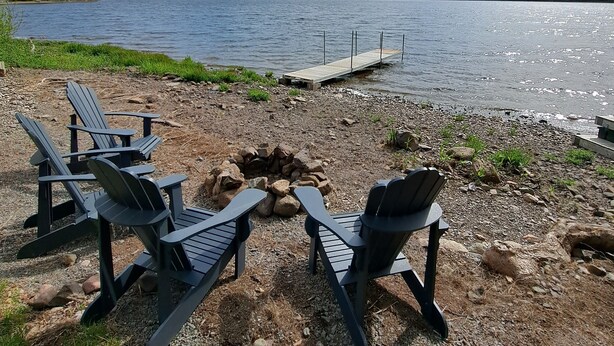 A lakeside setting with a wooden dock extending into the water, surrounded by a gravel area with several Adirondack chairs. - Lochaber Lake View Cottage (Antigonish County)