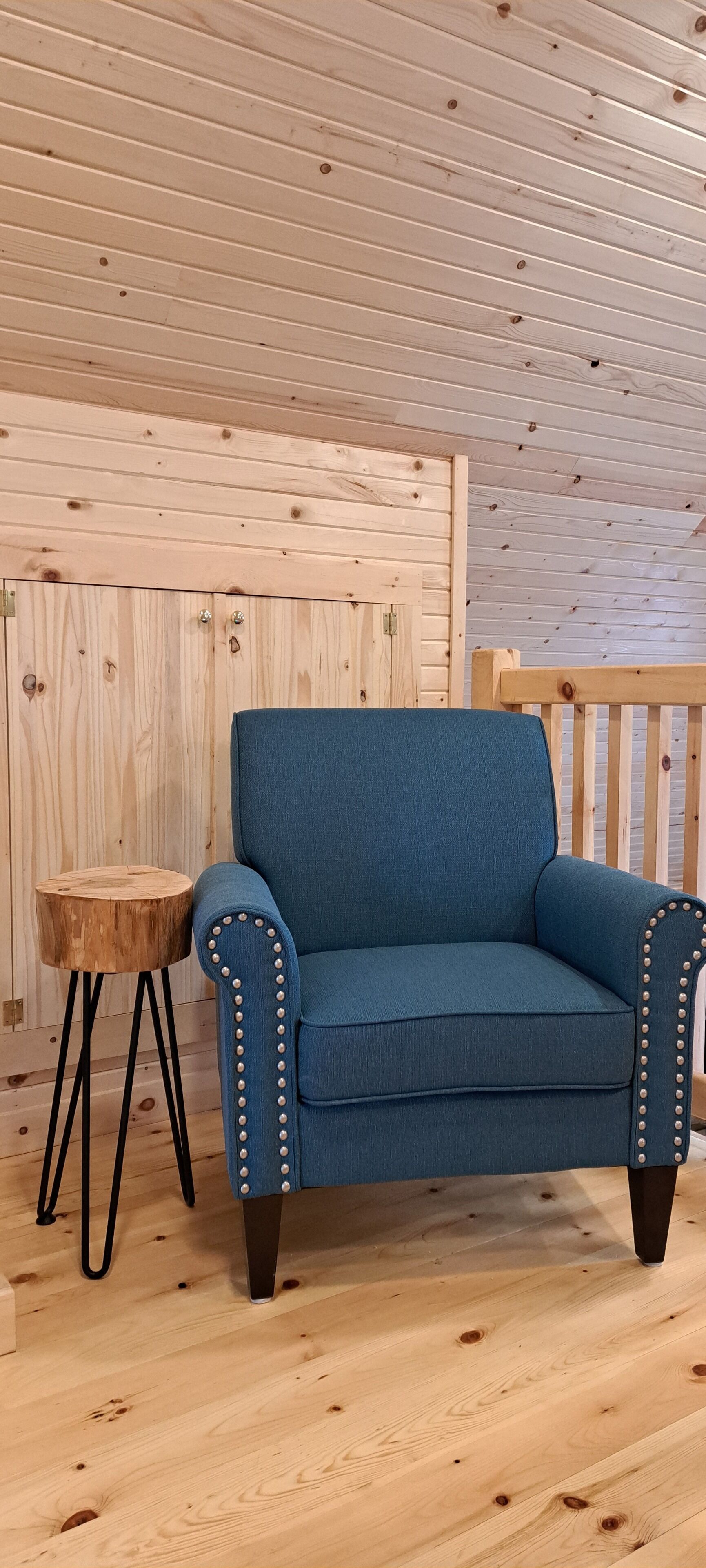 Un sillón azul con tachuelas metálicas, un taburete de madera y un suelo de madera en una habitación con paneles de madera en el techo.