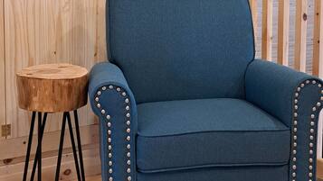 Un sillón azul con tachuelas metálicas, un taburete de madera y un suelo de madera en una habitación con paneles de madera en el techo.