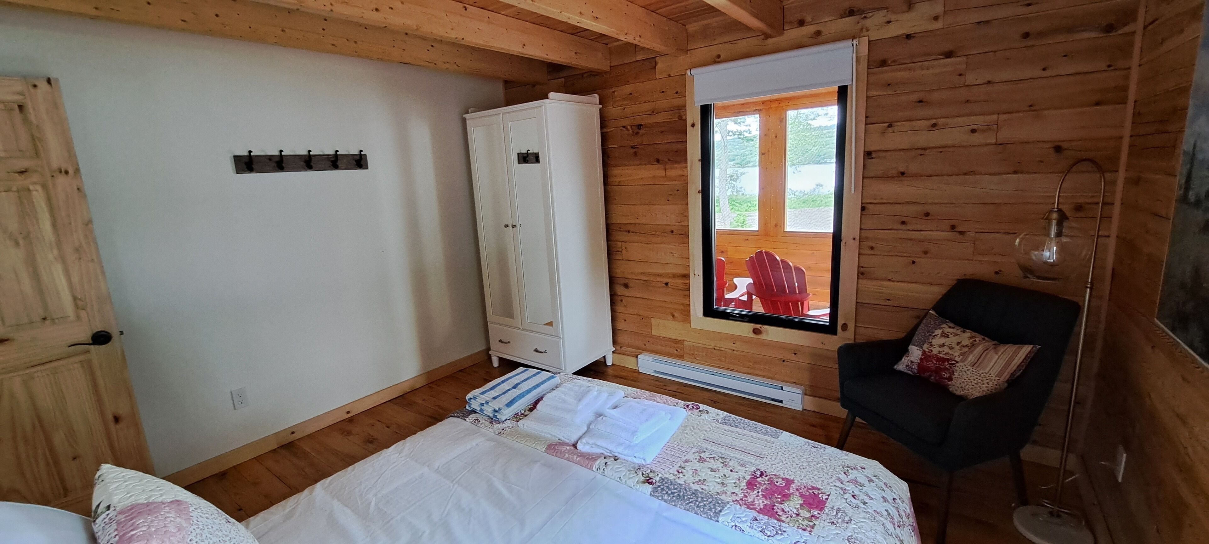 A wooden-paneled room with a bed, a white wardrobe, a red chair, and a window with a view of greenery.