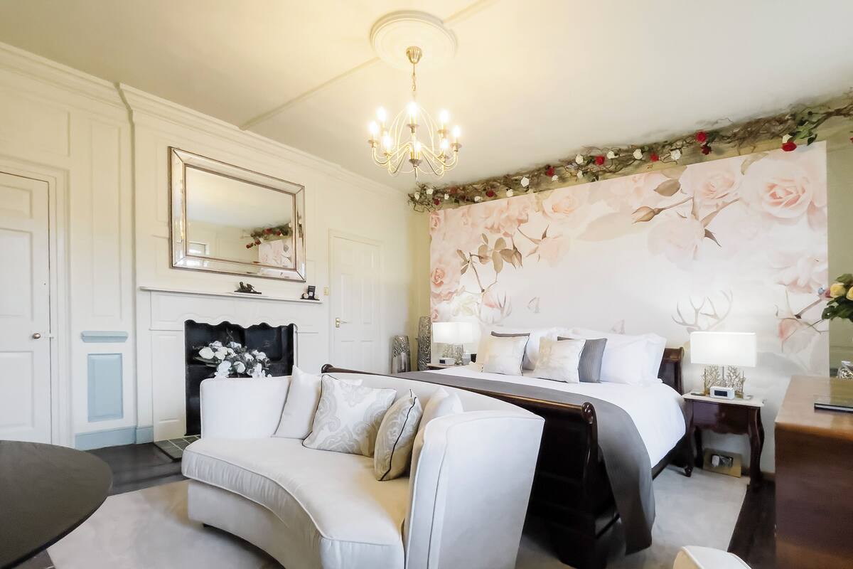 Een hotelkamer met een gebogen witte bank, een bed met witte beddengoed, een grote spiegel boven de open haard, en bloemenbehang.