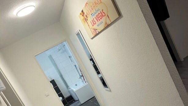 Un couloir avec un tapis gris, une pancarte d’accueil encadrée, et une salle de bain visible au fond.