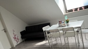 Une pièce moderne et bien éclairée, avec une table à manger, des chaises blanches, un canapé noir et une fenêtre avec des stores.