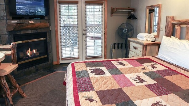 A cozy room with a fireplace, a large bed with a quilt, a television, and a sliding glass door. - Fawn Valley Inn: 254 1 Bedroom Apts (Estes Park)