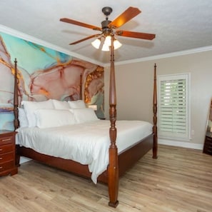 A four-poster bed with a ceiling fan, wooden floor, and a painting on the wall.
