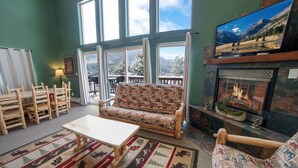 A cozy living room with a fireplace, wooden furniture, and large windows offering a view of the outdoors. - Lazy R Cottages: 16a 2 Bedroom Cabin (Estes Park)