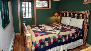 Un dormitorio con cabecera de madera, colcha, lámpara de noche y puerta con ventanal.