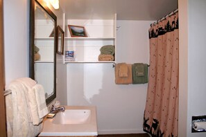 A bathroom with a white sink, a mirror, a towel rack with two towels, a shelf with a picture and a box, and a shower curtain with a forest pattern. - Lazy R Cottages: 2 2 Bedroom Cabin (Estes Park)