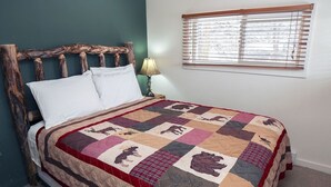 A bed with a quilt featuring animal prints, a wooden headboard, a bedside lamp, and a window with blinds. - Triple R Cottages: 2 1 Bedroom Cabin (Estes Park)