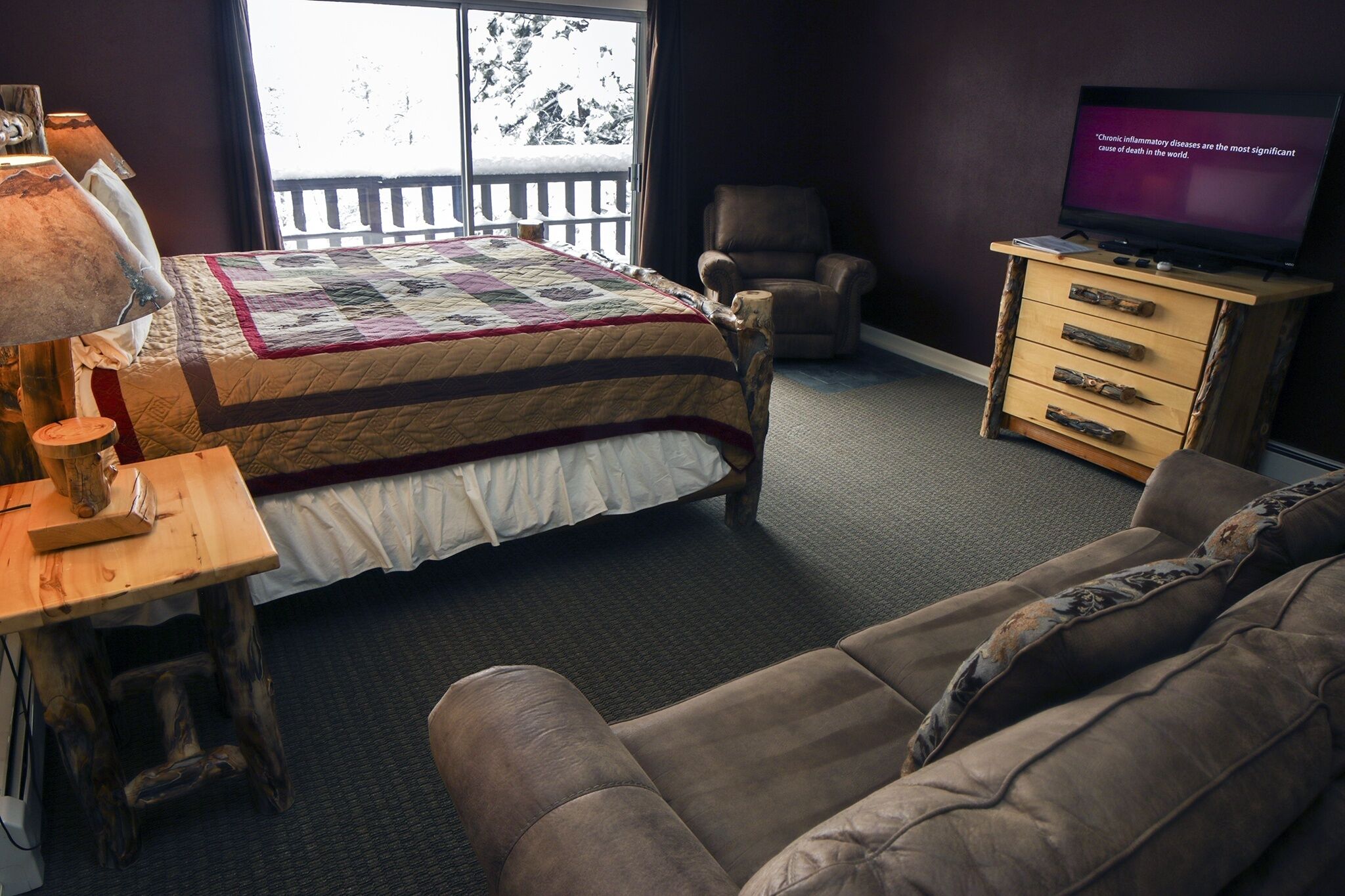 Quarto com cama, mesa de cabeceira de madeira, televisão, poltrona e sofá.