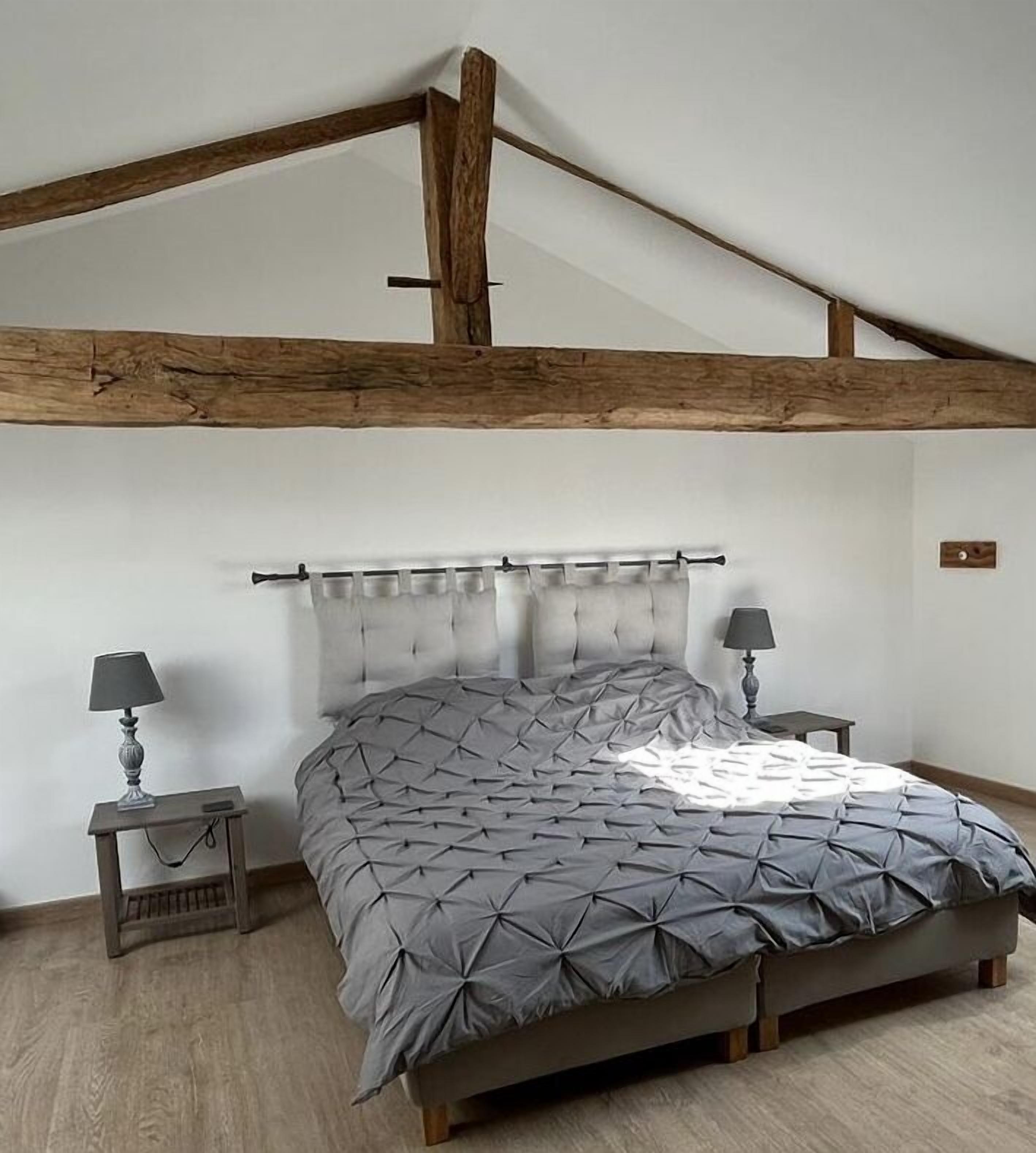 A bedroom with a sloped ceiling, exposed wooden beams, a tufted headboard, a grey patterned bedspread, two bedside tables with lamps, and a light-colored wooden floor.