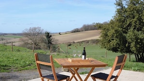 Ein Holztisch mit Stühlen steht auf einer Terrasse mit Blick auf ein grünes Feld und Bäume.
