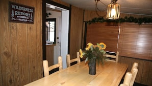 A wooden dining table with a vase of sunflowers, wooden chairs, and a sign reading "WILDERNESS RESORT ROUGHING IT IN STYLE." - Triple R Cottages: 1 3 Bedroom Cabin (Estes Park)