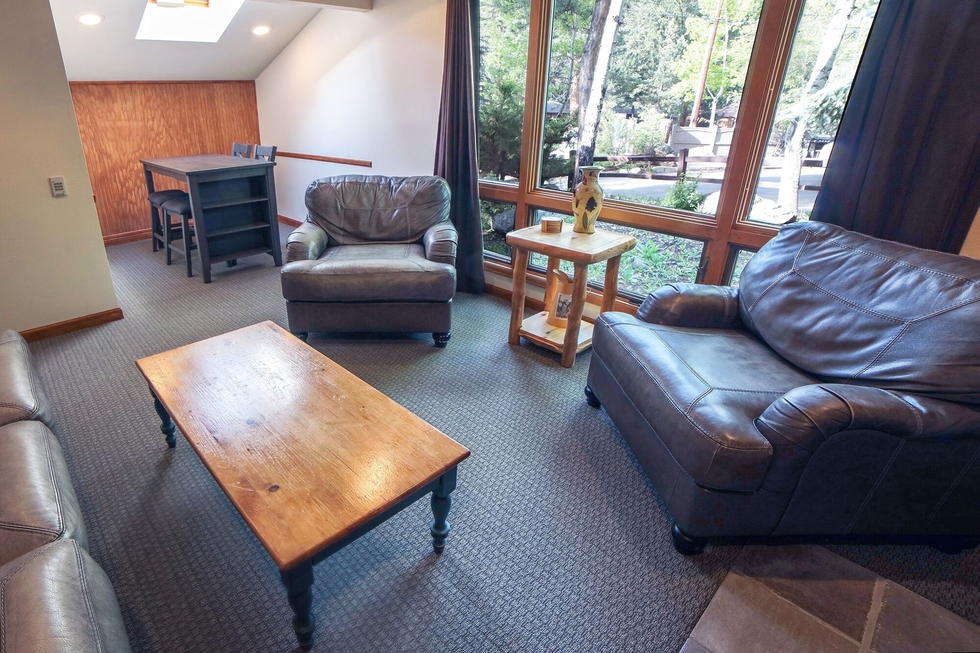 A living room with a leather sofa, a wooden coffee table, and a leather armchair. There is a small wooden table with a vase and a picture frame. The room has large windows with a view of trees and a building outside.