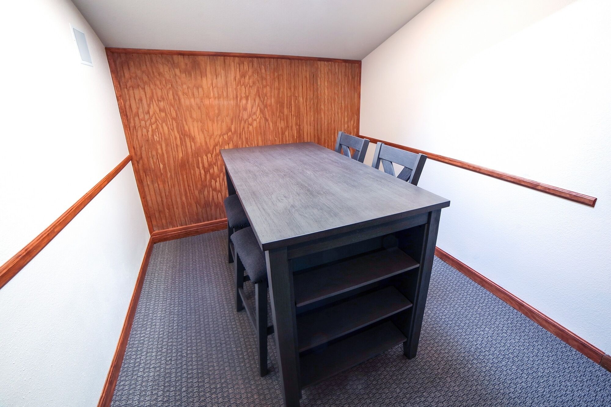 A small, rectangular room with a dark wooden table and four chairs. The room has a wooden panel wall and a carpeted floor.