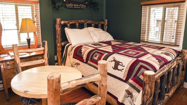 A rustic bedroom with a wooden bed, a round wooden table, and a lamp. - Triple R Cottages: 4 1 Bedroom Cabin (Estes Park)