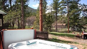 A hot tub in a backyard with a wooden deck, surrounded by tall pine trees and a mountain in the background. - Triple R Cottages: 8 2 Bedroom Cabin (Estes Park)