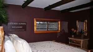 A bedroom with a bed, a window with blinds, a sign on the wall, and a dresser with a mirror and flowers. - Triple R Cottages: 8 2 Bedroom Cabin (Estes Park)