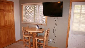 A small room with a round wooden table and four chairs, a television mounted on the wall, and a window with blinds. - Fawn Valley Inn: 253 1 Bedroom Apts (Estes Park)