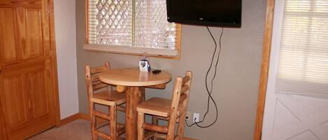 A small room with a round wooden table and four chairs, a television mounted on the wall, and a window with blinds.