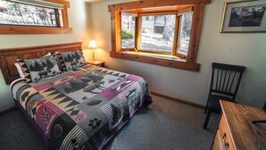 A bedroom with a bed, a nightstand, a lamp, a chair, a desk, a window with blinds, and a ceiling fan. - Sunnyside Knoll: 8 1 Bedroom Cabin (Estes Park)