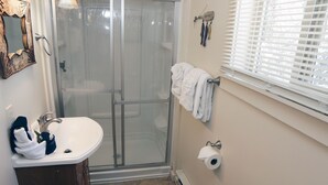 Et badeværelse med glasbrusekabine, en hvid vask, et håndklædestativ med håndklæder, toilet og et vindue med persienner.
