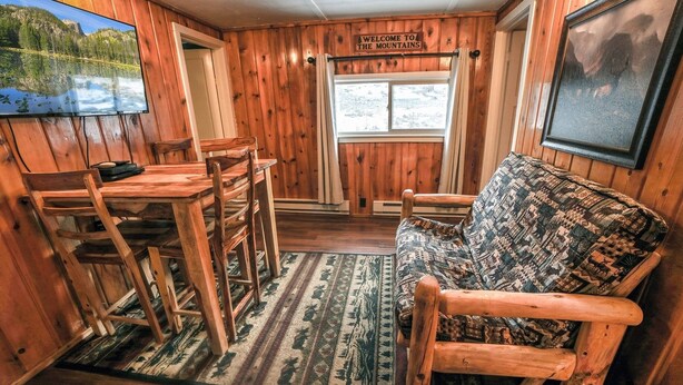 Et interiør af en træhytte med et spisebord, stole, en stribet sofa og et vindue med et skilt, der siger "Welcome to the Mountains".