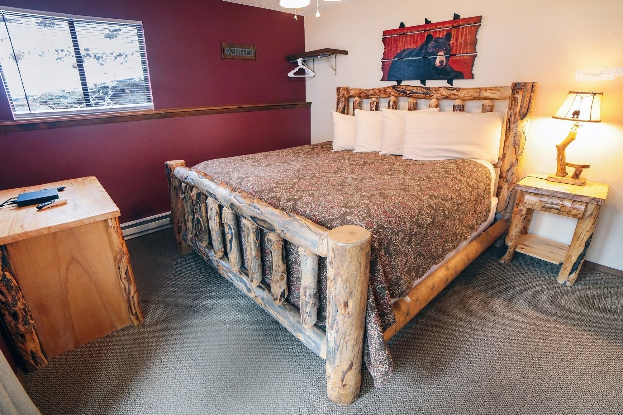 Une chambre à coucher rustique avec un cadre de lit en bois, un panneau d’accueil, un tableau représentant un ours et une lampe sur une table de chevet.
