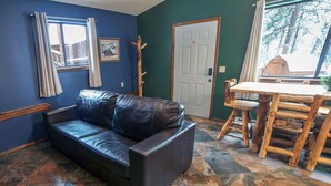 Una habitación con un sofá de cuero negro, una mesa de comedor de madera con sillas, una puerta y una ventana con cortinas.