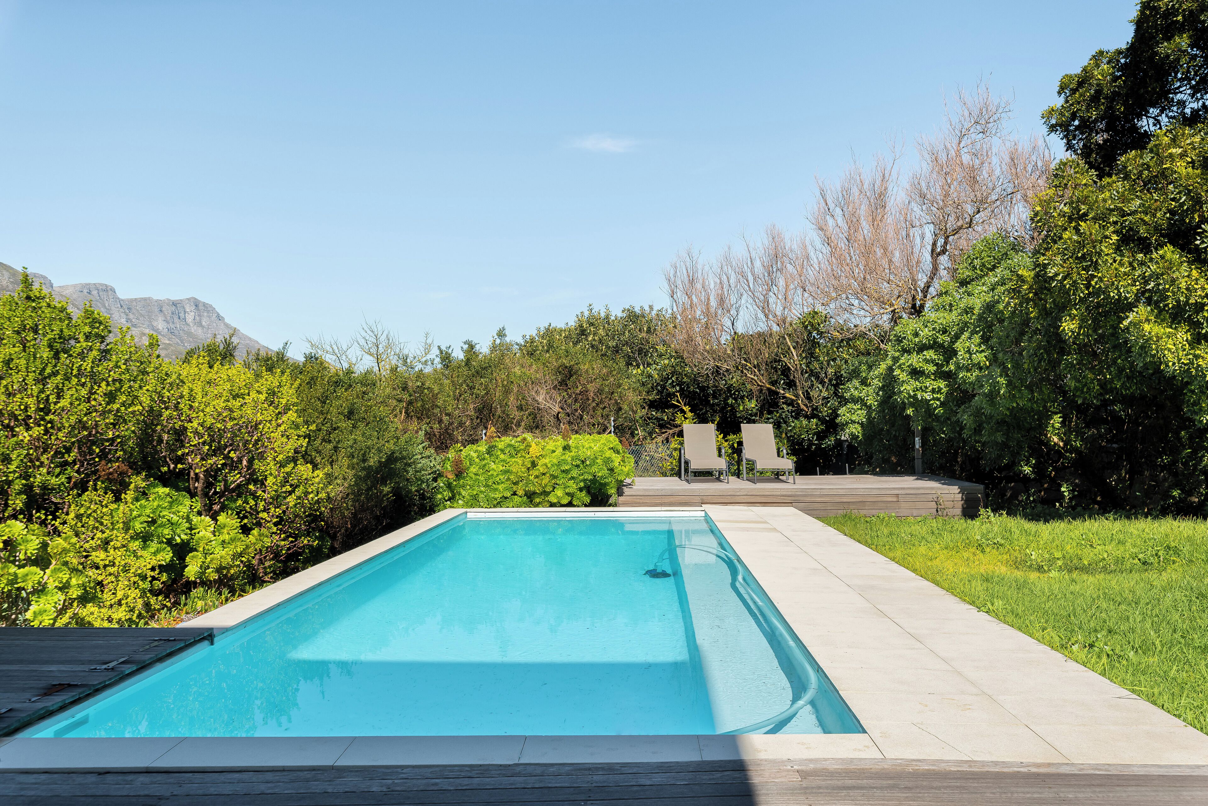A rectangular swimming pool with a wooden deck surrounding it, set against a backdrop of lush greenery and mountains.
