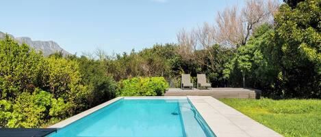 A rectangular swimming pool with a wooden deck surrounding it, set against a backdrop of lush greenery and mountains.