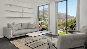 Ein modernes Wohnzimmer mit einem grauen Sofa, einem Couchtisch und Blick auf Berge und Grün durch große Fenster.