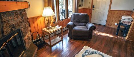 Una sala de estar acogedora con chimenea de piedra, un sillón cómodo, una mesa de centro de madera y un ventanal con vistas a árboles.
