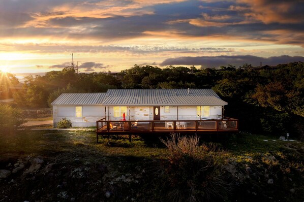 A white house with a wooden deck, surrounded by trees and a sunset in the background.