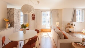 A modern living room with a white sofa, a dining table with chairs, and a large window.