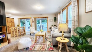 A cozy living room with a wooden dining table, a sofa, and a coffee table. There are large windows with curtains, a TV, and a bookshelf. - Cozy cabin on 5 acres of wooded land! (Wausaukee)