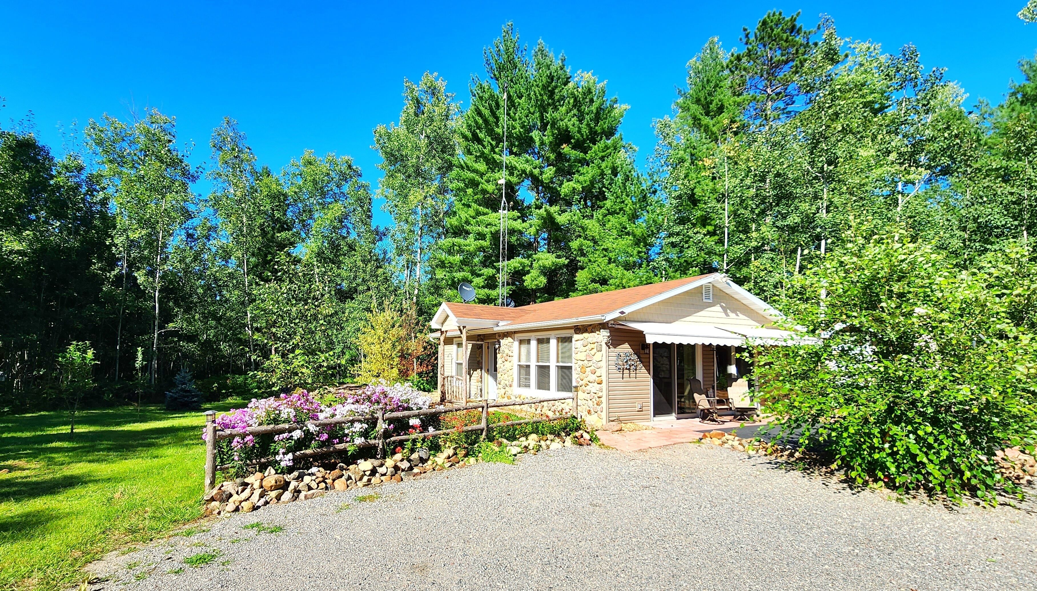 A small stone cabin with a porch, surrounded by a gravel driveway and a flower bed.