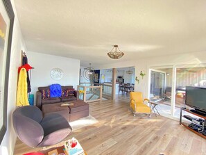 A spacious living room with a sofa, a yellow armchair, and a television. A kitchen is visible in the background, and there is a large window providing natural light. - Luxe Lakefront, King & Queen Bed Suites, Sunset, Pools, Boat Slip, 1,786 sqft! (Osage Beach)