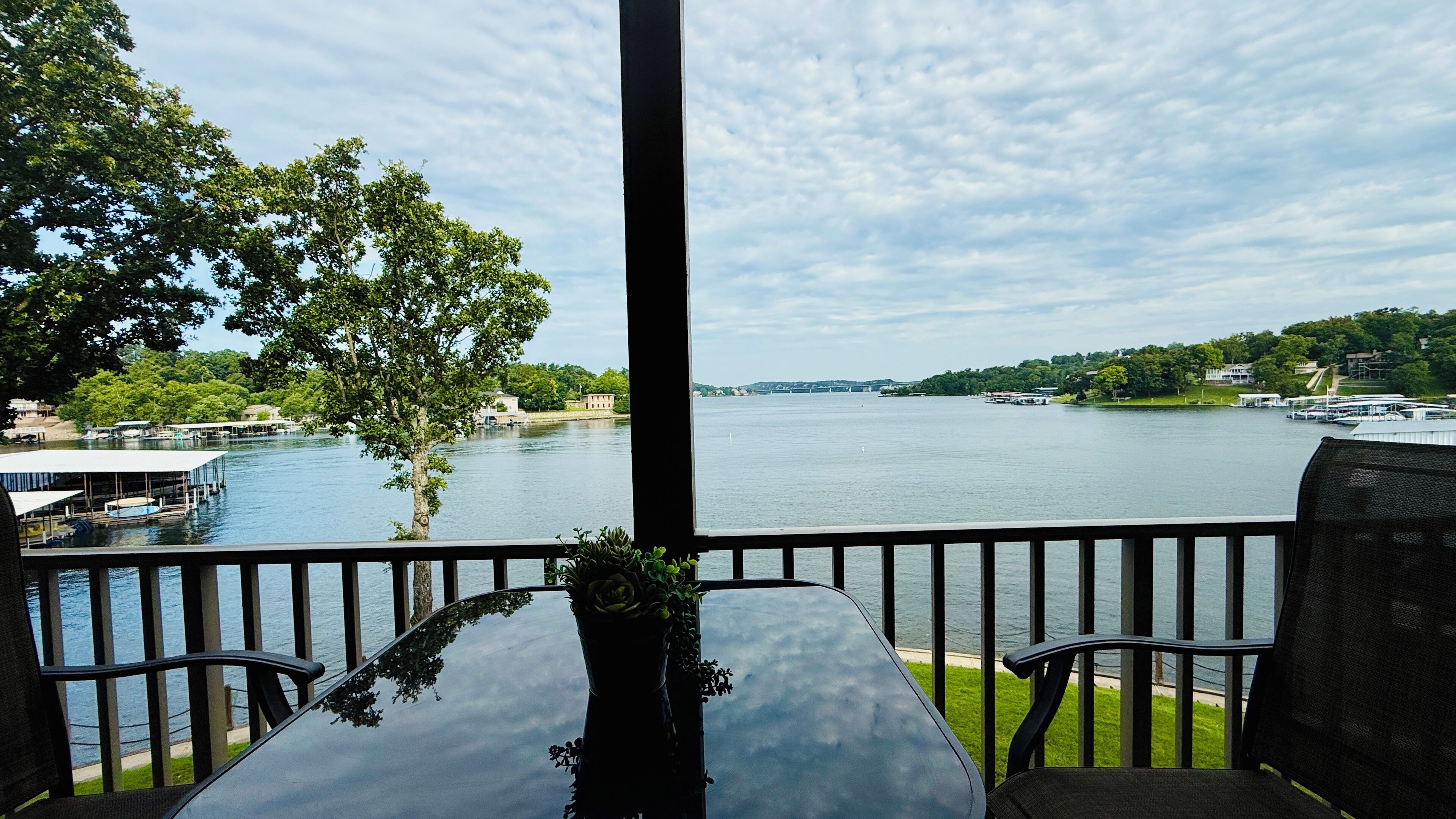 A terrace with a table and chairs overlooking a river, with boats and buildings in the background.