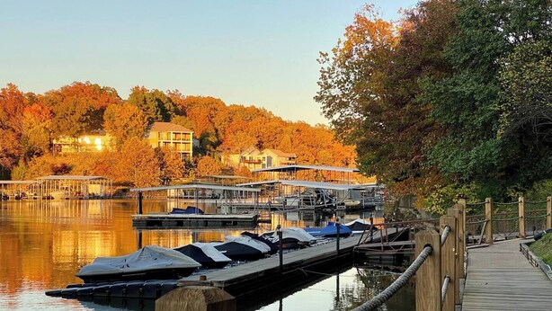 A wooden dock extending into calm water with boats docked, surrounded by autumn-colored trees and buildings in the background. - Luxe Lakefront, King & Queen Bed Suites, Sunset, Pools, Boat Slip, 1,786 sqft! (Osage Beach)