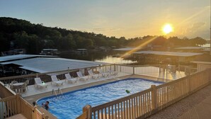 A pool area with a view of the sunset over a body of water, surrounded by a wooden deck and lounge chairs. - Luxe Lakefront, King & Queen Bed Suites, Sunset, Pools, Boat Slip, 1,786 sqft! (Osage Beach)