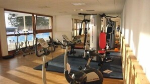 A gym with cardio machines, weight equipment, and a large window with a view of trees. - Le Silène - Appt pour 6 (Chamrousse)