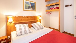 A bedroom with a wooden headboard, two bedside lamps, a bed with striped pillows, a red blanket, a framed picture of a landscape, and a shelf with folded clothes. - Le Silène - Appt pour 6 (Chamrousse)