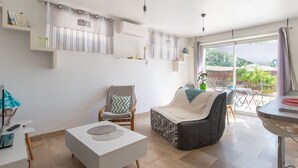 Un salon moderne avec un canapé blanc, un fauteuil gris et une table basse blanche. On y trouve une table à manger avec des chaises, une grande fenêtre avec des rideaux et une vue sur la verdure à l’extérieur.