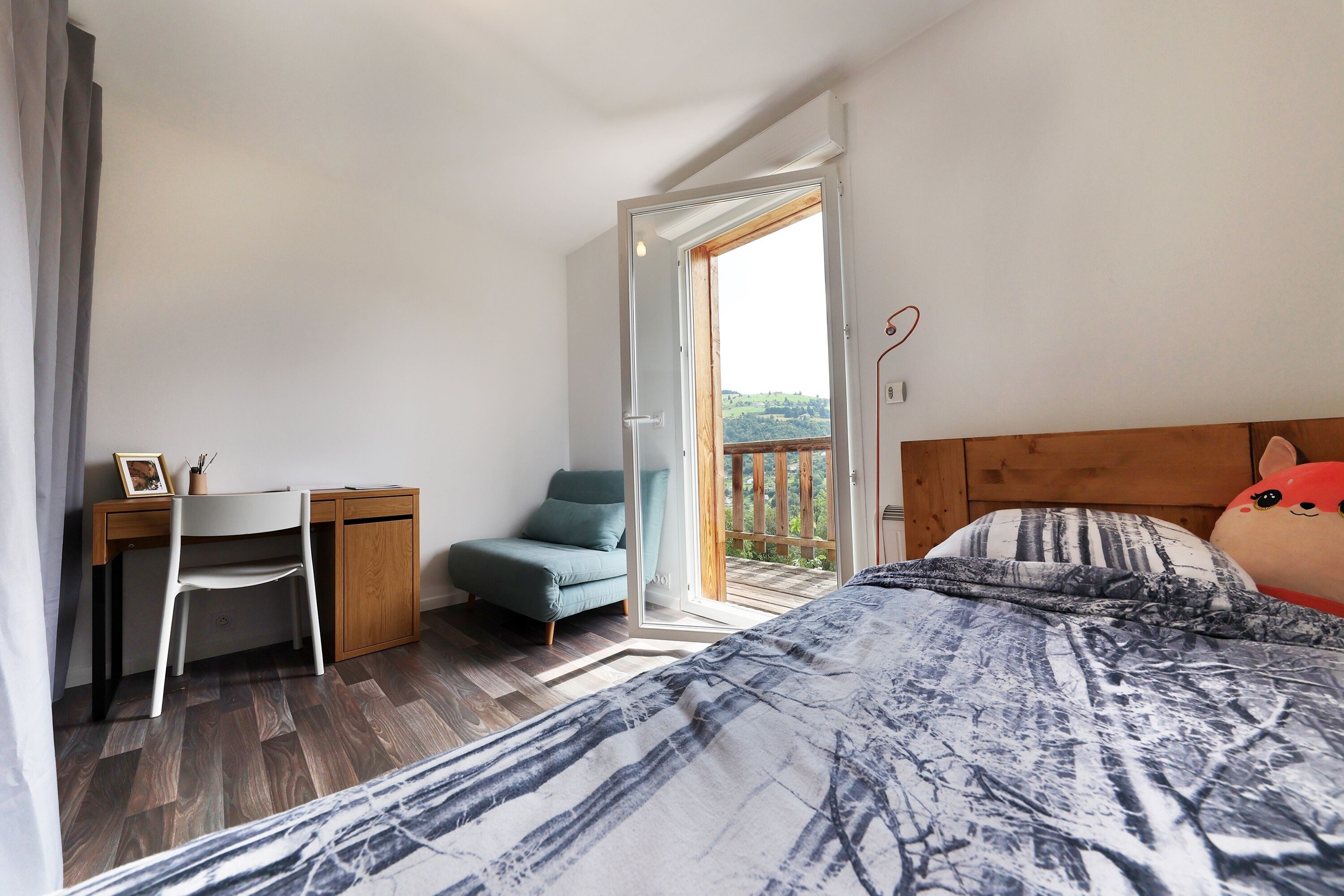 A bedroom with a bed, a desk, a chair, and a balcony with a view.