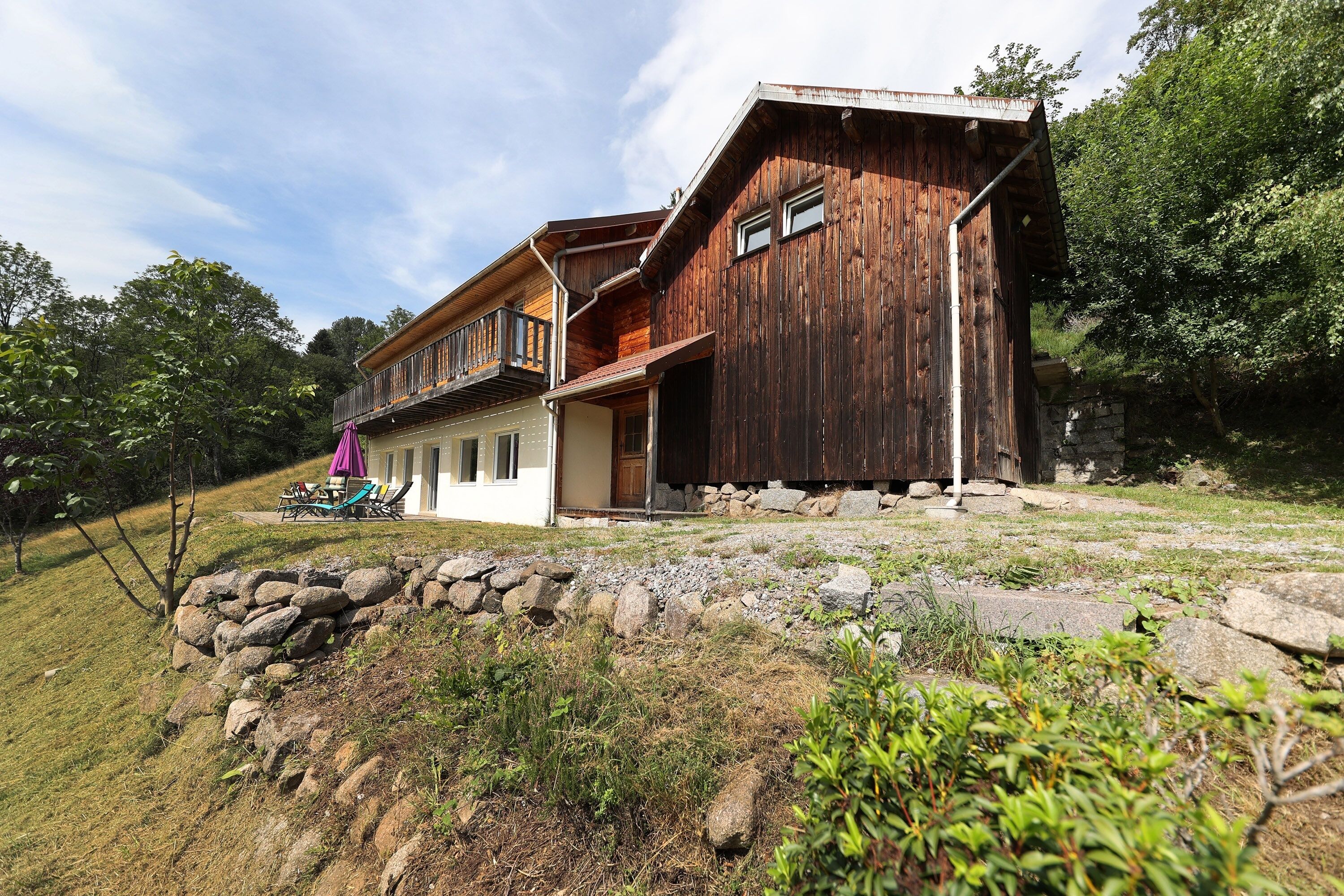 Un chalet rustique en bois doté d’un balcon, entouré d’un mur de pierres et de verdure.
