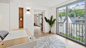 A modern hotel room with a large window, a wooden desk, a potted plant, and a view of the cityscape. - Modern Apt Old Town w Panoramic View ID4 (Luxembourg City)
