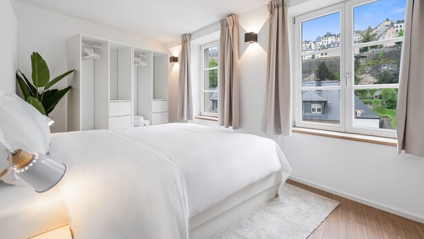 A bedroom with a large bed, a wardrobe, a mirror, and a window with a view of buildings. - Modern Apt Old Town w Panoramic View ID4 (Luxembourg City)