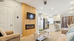 A modern living room with a fireplace, a television, and a kitchen area in the background.